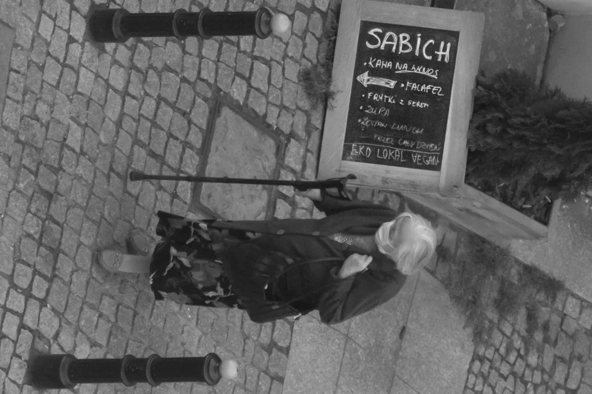 Elderly woman with a cane standing beside a vegan cafe sign seen from above